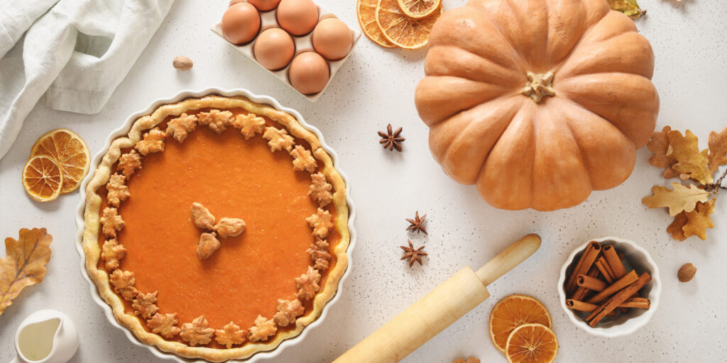 Imagine de sus placinta de dovleac, asezata pe un blat de bucatarie alb. Lângă e un facalet, un bol cu bețișoare de scorțișoară, felii uscate de portocale, steluțe de anason și un dovleac. Decor de toamnă