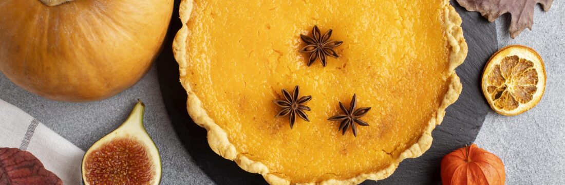 Imagine de sus, plăcintă de dovleac simplă decorată cu steluțe de anason. Lângă, un dovleac, jumătate de smochină, tacâmuri, decor de toamnă