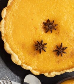 Imagine de sus, plăcintă de dovleac simplă decorată cu steluțe de anason. Lângă, un dovleac, jumătate de smochină, tacâmuri, decor de toamnă