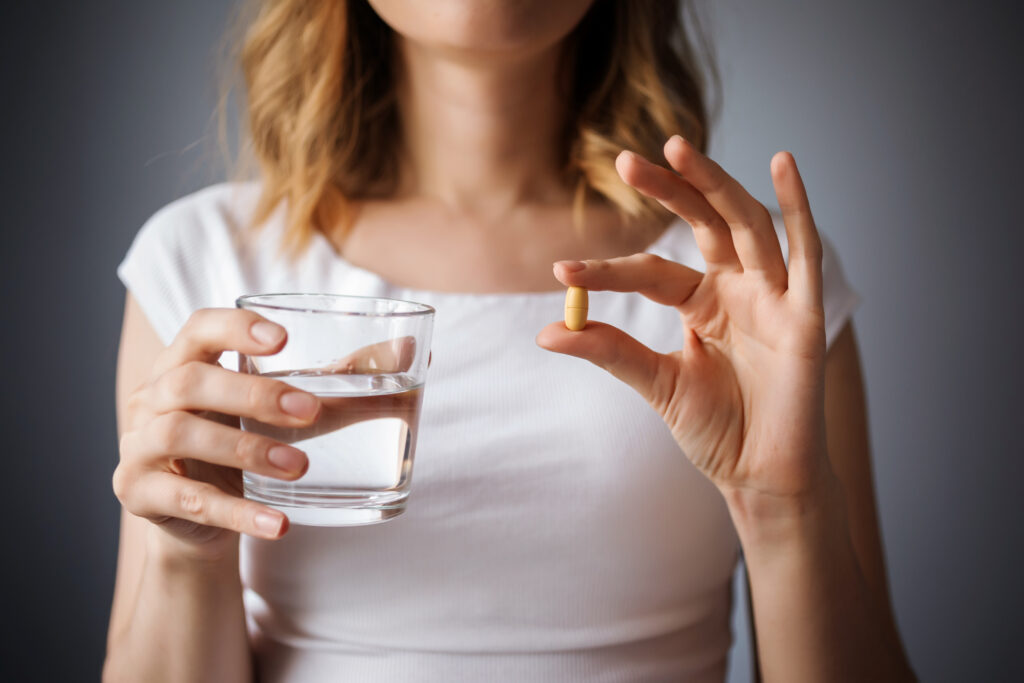close up cu pieptul unei femei, intr-o mana are o pastila, supliment alimentar, in alta un pahar un pahar cu apa