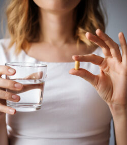 close up cu pieptul unei femei, intr-o mana are o pastila, supliment alimentar, in alta un pahar un pahar cu apa