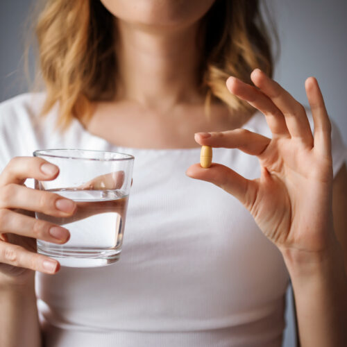 close up cu pieptul unei femei, intr-o mana are o pastila, supliment alimentar, in alta un pahar un pahar cu apa