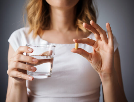 close up cu pieptul unei femei, intr-o mana are o pastila, supliment alimentar, in alta un pahar un pahar cu apa